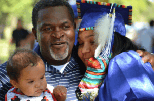 A NICC student in a graduation cap and gown embraces her partner and child.