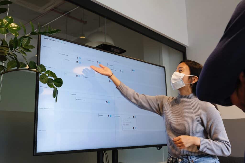 A woman wearing a face mask gestures to a large display screen.