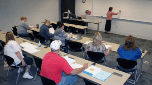 Multiple people sit in a small classroom as a woman writes on a whiteboard at the front of the room.