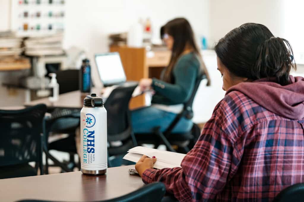 A young woman facing away from the camera reads a textbook. A water bottle with the NHS College logo sits beside her. Another woman works on a laptop in the background.