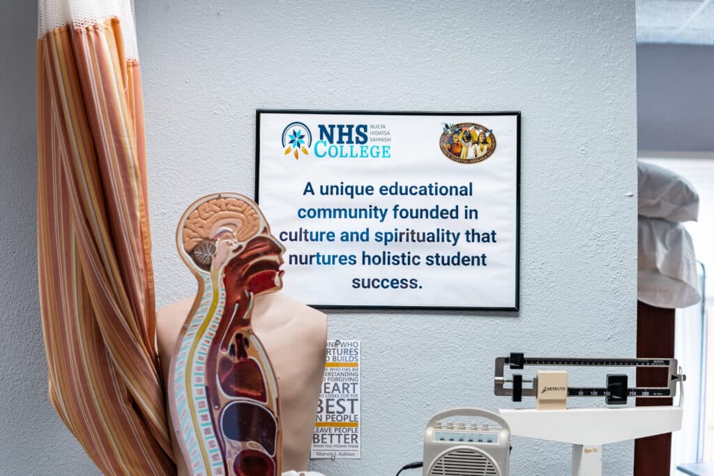 An anatomical model and various medical training equipment stand in front of a poster that reads: "NHS College: A unique educational community founded in culture and spirituality that nurtures holistic student success."