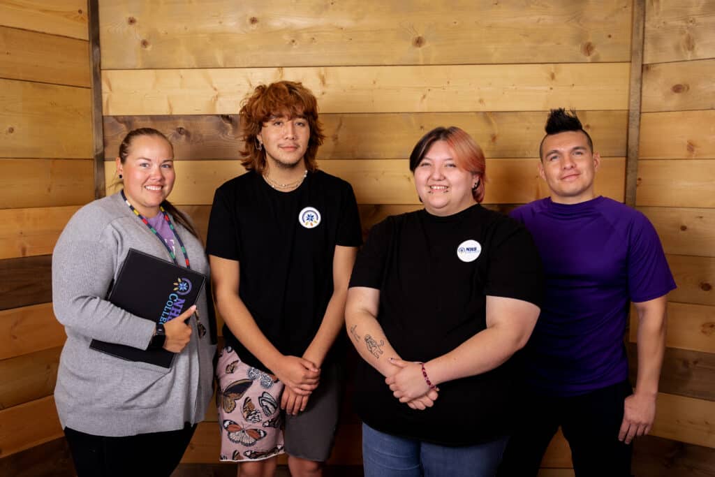 Four people stand in a row in front of wood paneling and smile at the camera.