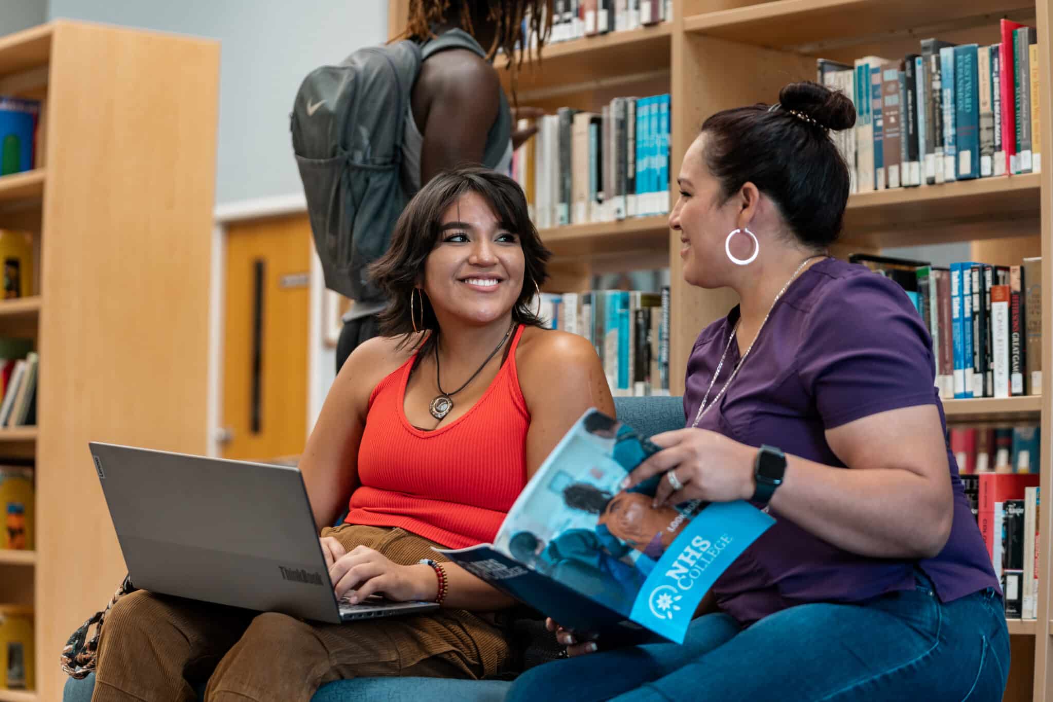 Two women sit in a library setting and are smiling at each other. A man browses bookshelves in the background.